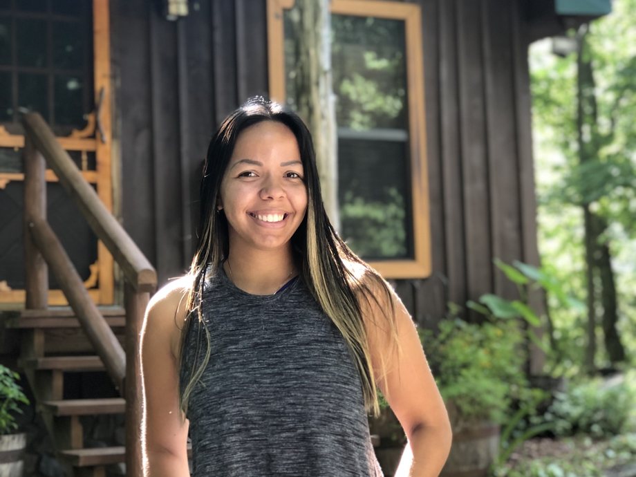 Anahely sonriendo frente a una cabaña de madera en el bosque